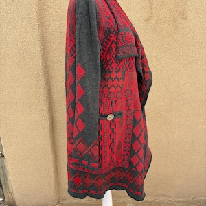 Puede incluir: Un c&aacute;rdigan estampado en rojo y gris con un dise&ntilde;o geom&eacute;trico. El su&eacute;ter de manga larga presenta un cierre de bot&oacute;n y un cuello chal. El c&aacute;rdigan se exhibe en un maniqu&iacute; blanco.