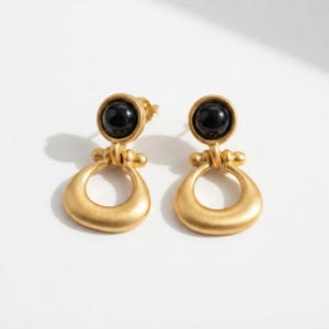May include: A pair of drop earrings featuring a matte gold finish. Each earring consists of a circular black stone set within a gold bezel, connected to a larger, open-work gold hoop shaped like a rounded horseshoe or a stylized teardrop link.