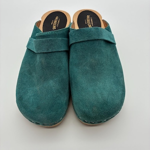May include: A pair of teal suede clogs with wooden soles and a wide strap across the top. The black insoles feature the text Swedish Hasbeens printed in white. The shoes have a rounded toe design and visible stitching along the wooden base.