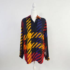 May include: A long-sleeved button-down shirt with a vibrant geometric pattern. The shirt features a black base with bold stripes and houndstooth-like shapes in yellow, red, and blue. The collar and cuffs have a contrasting striped design.