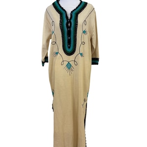 May include: A beige, long-sleeved dress with a V-neckline and a floor-length hem. The dress features teal and black embroidered detailing around the neckline, sleeves, and hem. The dress is displayed on a mannequin.