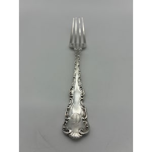 May include: A silver-toned metal fork featuring an ornate, embossed scrollwork pattern along the handle. The base of the handle displays a prominent, engraved capital letter D in a decorative script font. The fork has four slender tines.