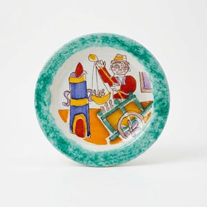 May include: A hand-painted ceramic plate with a green speckled rim. The plate features a colorful illustration of a person weighing items, a candle, and a cart. The artwork uses bright colors like blue, red, yellow, and orange.
