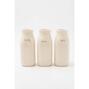 May include: Three off-white ceramic milk bottles with a smooth finish. Each bottle features a narrow neck and a rounded base. The first bottle is inscribed with the word beauty., the second with peace., and the third with the word grow. in black.