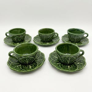May include: Five green ceramic teacups and saucers. The cups and saucers are designed to resemble cabbage leaves. The teacups have a small handle and the saucers are round. The set is placed on a white surface.