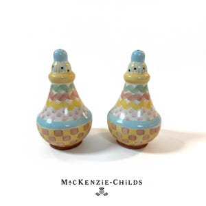 May include: Two decorative salt and pepper shakers with a colorful, whimsical design. The shakers feature a light blue top, a yellow and pastel-colored body with a checkered pattern at the base. The brand name "MacKenzie-Childs" is printed at the bottom.