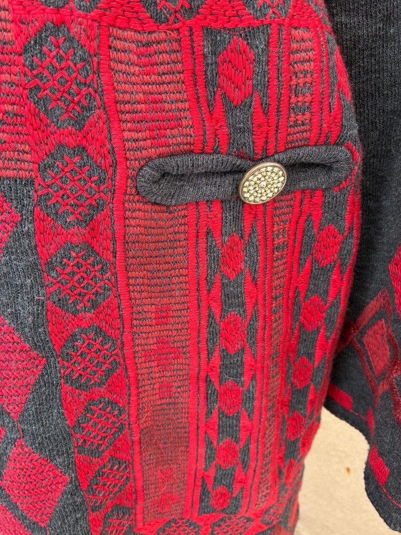 Puede incluir: Primer plano de una chaqueta estampada roja y gris. La chaqueta presenta intrincados dise&ntilde;os bordados en rojo sobre un fondo gris. Se ve un peque&ntilde;o bolsillo con un bot&oacute;n decorativo.