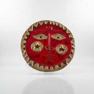 May include: A red ceramic plate with a sun face design. The plate features a red background with gold-colored eyes, star-shaped cheeks, and a decorative border. The plate is displayed on a clear stand.