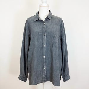 May include: A gray button-down shirt with long sleeves and a single chest pocket. The shirt is displayed on a white mannequin against a white background. The shirt has white buttons and a classic collar.