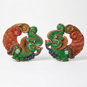 May include: A pair of hand-carved wooden sculptures depicting mythical creatures with green bodies, red and orange curved tails, and blue accents. Each figure features intricate scale patterns, a wide open mouth with visible teeth, and a circular design.