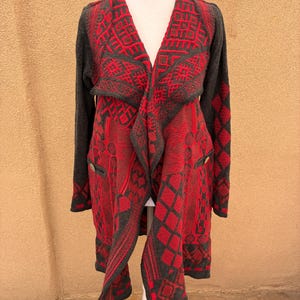 Puede incluir: Un c&aacute;rdigan largo y abierto en rojo y gris, exhibido en un maniqu&iacute;. El su&eacute;ter presenta un patr&oacute;n geom&eacute;trico y dos peque&ntilde;os botones. Las mangas y el ribete son grises, mientras que el cuerpo es predominantemente rojo.