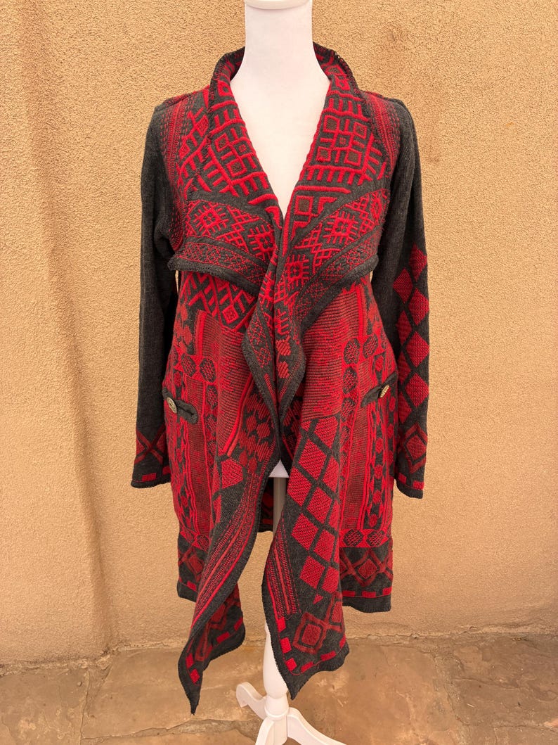 Puede incluir: Un c&aacute;rdigan largo y abierto en rojo y gris, exhibido en un maniqu&iacute;. El su&eacute;ter presenta un patr&oacute;n geom&eacute;trico y dos peque&ntilde;os botones. Las mangas y el ribete son grises, mientras que el cuerpo es predominantemente rojo.