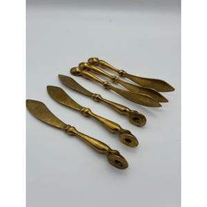 May include: A set of five brass butter spreaders with a metallic gold finish. Each spreader features a flat, pointed blade and a rounded, decorative handle with a small circular indentation at the end, arranged in a diagonal pattern.