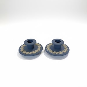 May include: Two blue bottle stoppers with a circular design and a beige leaf pattern around the edge. The stoppers have a cylindrical top and a flat, round base. The background is white.