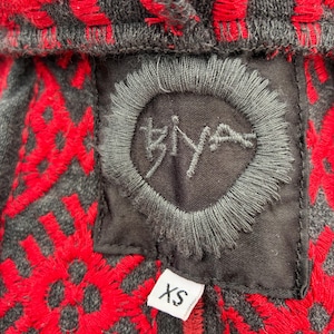Puede incluir: Primer plano de una sudadera con capucha gris con patrones geom&eacute;tricos bordados en rojo. La sudadera con capucha presenta un parche negro con la palabra "Biya" bordada en un c&iacute;rculo. Una etiqueta blanca con "XS" est&aacute; adjunta.