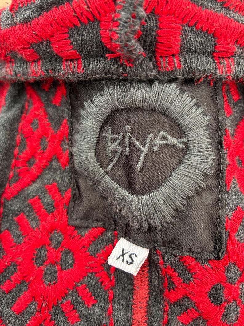 Puede incluir: Primer plano de una sudadera con capucha gris con patrones geom&eacute;tricos bordados en rojo. La sudadera con capucha presenta un parche negro con la palabra "Biya" bordada en un c&iacute;rculo. Una etiqueta blanca con "XS" est&aacute; adjunta.