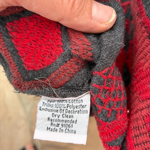 Puede incluir: Primer plano de una tela estampada en rojo y gris. La tela presenta un dise&ntilde;o geom&eacute;trico con formas de diamante rojas y bordes grises. Una etiqueta blanca con texto es visible, que indica la composici&oacute;n del material y las instrucciones de cuidado.