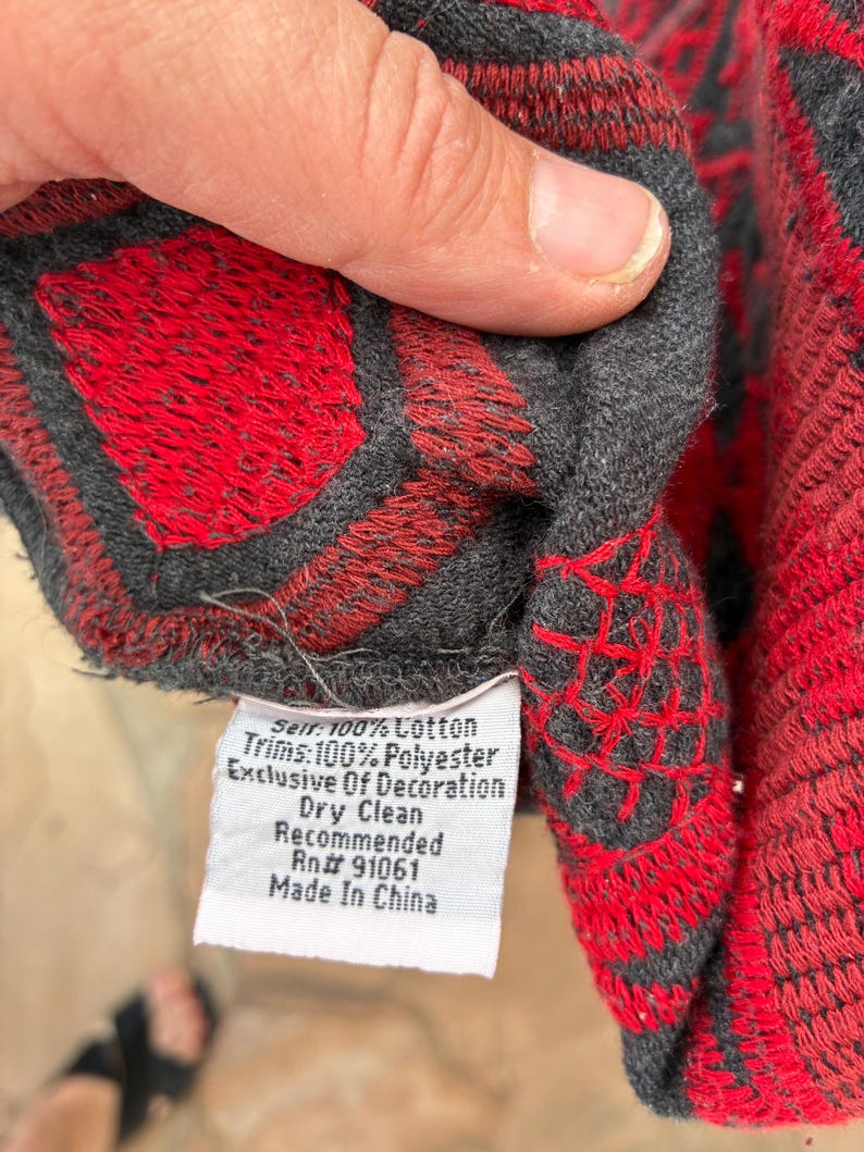 Puede incluir: Primer plano de una tela estampada en rojo y gris. La tela presenta un dise&ntilde;o geom&eacute;trico con formas de diamante rojas y bordes grises. Una etiqueta blanca con texto es visible, que indica la composici&oacute;n del material y las instrucciones de cuidado.