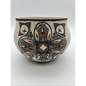May include: A rounded ceramic bowl with a cream-colored base featuring a hand-painted geometric design in black and terracotta brown. The pattern consists of symmetrical floral-like shapes with cross-hatched details and a solid black rim.