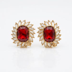 May include: A pair of gold-tone earrings featuring a large, rectangular, deep red center stone surrounded by a sunburst halo of clear baguette-cut crystals. Each earring includes a small gold-tone CD logo emblem at the bottom edge of the setting.