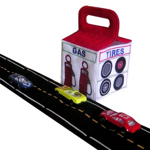 May include: A toy scene featuring a black road with yellow lane markings and three toy cars. A white and red gas station with the words "GAS" and "TIRES" is in the background.