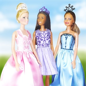 Puede incluir: Tres muñecas con vestidos de princesa y tiaras. La muñeca de la izquierda lleva un vestido rosa con ribetes de encaje blanco. La muñeca del centro lleva un vestido lavanda con bordados morados. La muñeca de la derecha lleva un vestido azul claro con ribetes negros.