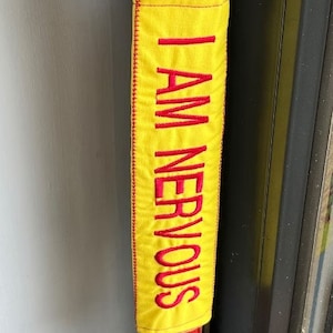 May include: A yellow fabric strap with red embroidered text that reads "I AM NERVOUS".