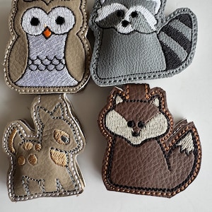 May include: Four embroidered animal patches: an owl, a raccoon, a deer, and a fox. The owl is brown and tan with an orange beak. The raccoon is grey with a striped tail. The deer is tan with brown spots. The fox is brown with a white face.