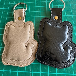 May include: Two cat-shaped keychains. One is tan with a stitched border, and the other is black with a shiny finish. Each keychain has a metal ring and a snap closure. The keychains are on a green cutting mat.