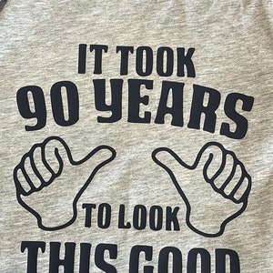 May include: Gray regular fit t-shirt with the text "IT TOOK 90 YEARS TO LOOK THIS GOOD" in navy blue. The design includes two thumbs-up gestures. The shirt has a crew neck and a tag.