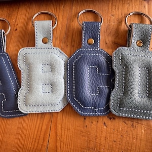 Op de afbeelding: Vier leren sleutelhangers met de letters A, B, C en D. Elke letter heeft een andere kleur: marineblauw, lichtblauw, donkerblauw en grijsgroen. De letters zijn gestikt met wit garen en bevestigd aan een metalen sleutelhanger.