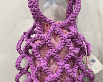 May include: A purple crocheted handbag featuring a thick, braided lattice exterior and a solid pink fabric drawstring pouch inside. A small white rectangular tag attached to the side displays the text CROCHET BY BEA in blue lettering.