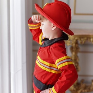 May include: A young child dressed as a firefighter, wearing a red helmet and a red and yellow fire-resistant suit, salutes with their right hand.