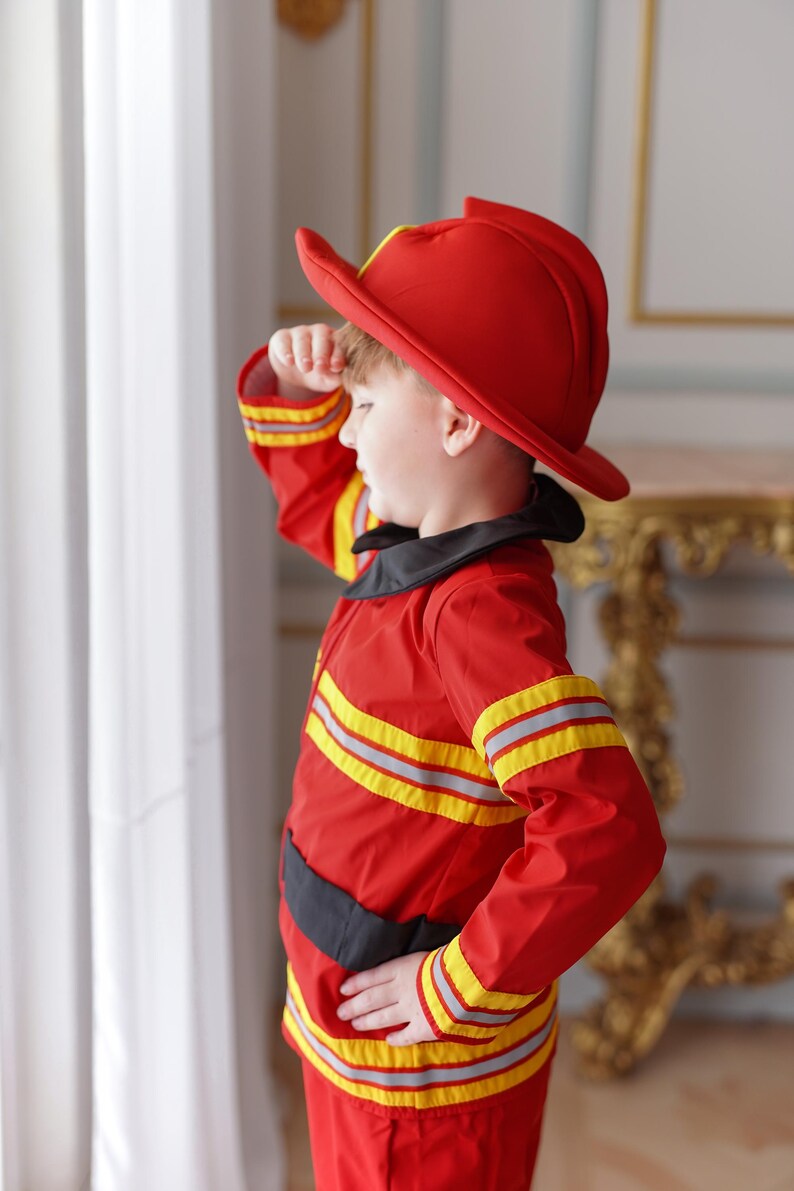 May include: A young child dressed as a firefighter, wearing a red helmet and a red and yellow fire-resistant suit, salutes with their right hand.