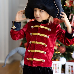 May include: A young child dressed as a toy soldier for the holidays. They are wearing a red velvet jacket with gold trim and buttons, black pants, and a black hat with gold trim. The child is standing in front of a Christmas tree.