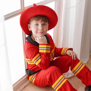 May include: A young boy dressed as a firefighter, wearing a red hat and red and yellow jumpsuit, sits by a window. He is looking at the camera and has his hand on his chin.