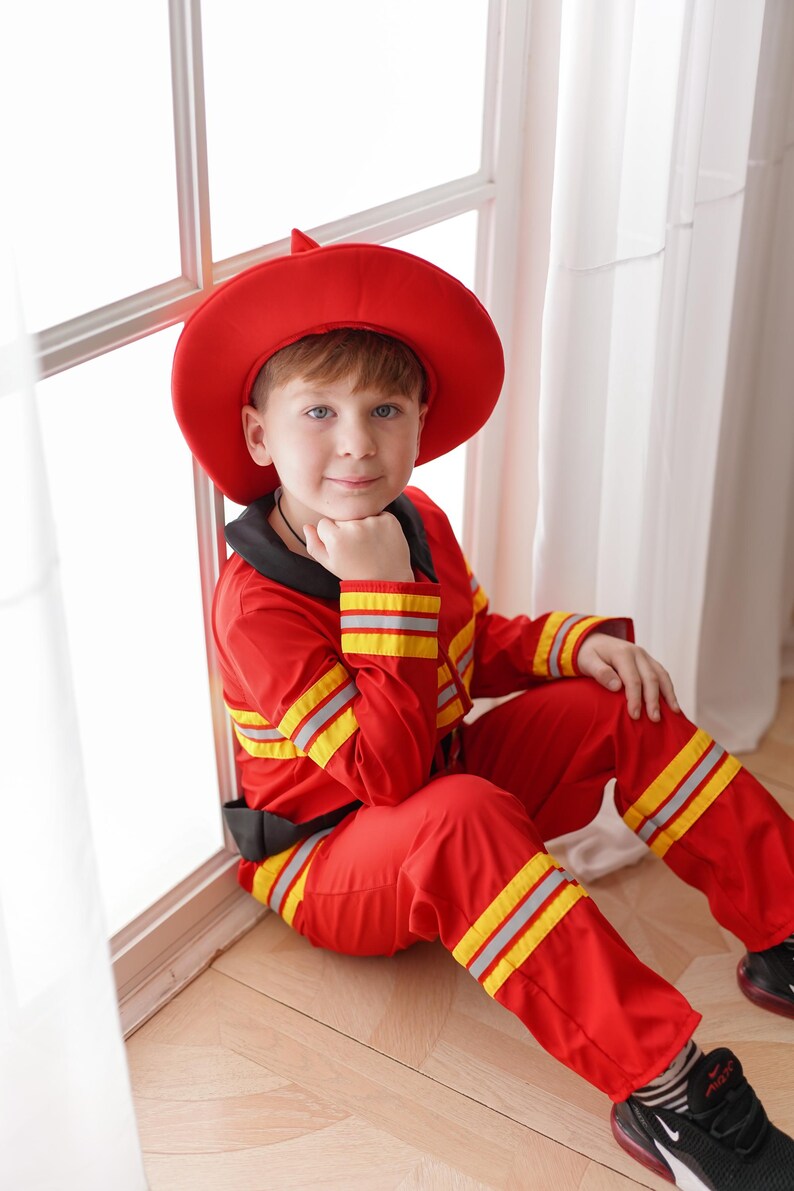 May include: A young boy dressed as a firefighter, wearing a red hat and red and yellow jumpsuit, sits by a window. He is looking at the camera and has his hand on his chin.