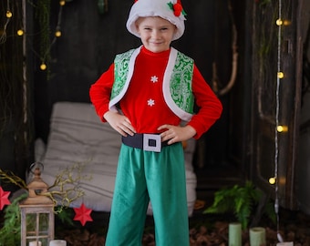 Puede incluir: Un disfraz festivo de elfo que incluye una camiseta roja de manga larga con copos de nieve blancos, un chaleco verde con ribete blanco, pantalones verdes, un cinturón negro con hebilla plateada, un gorro rojo y blanco y zapatos de elfo.