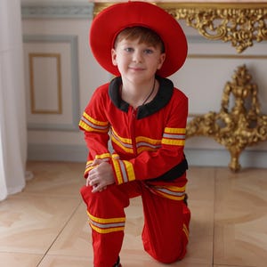 May include: A young boy dressed as a firefighter, wearing a red hat and a red and yellow striped jumpsuit, kneeling on a wood floor.