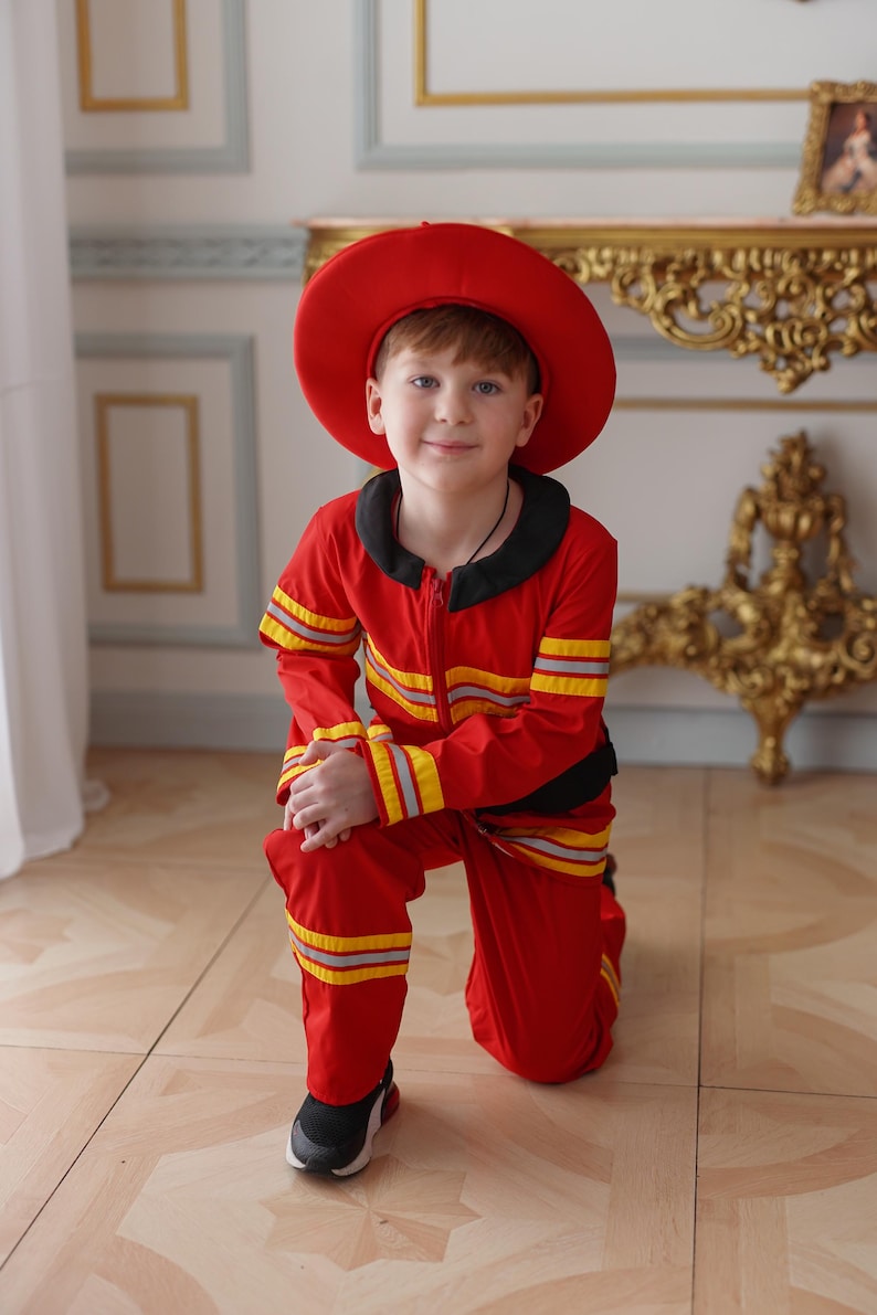 May include: A young boy dressed as a firefighter, wearing a red hat and a red and yellow striped jumpsuit, kneeling on a wood floor.