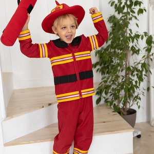May include: A young child dressed as a firefighter, wearing a red fire suit with yellow reflective stripes, a red firefighter hat with a yellow flame, and holding a red fire hose. The child is standing on a wooden staircase.