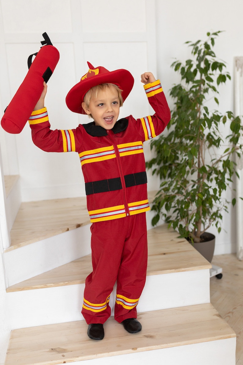 May include: A young child dressed as a firefighter, wearing a red fire suit with yellow reflective stripes, a red firefighter hat with a yellow flame, and holding a red fire hose. The child is standing on a wooden staircase.