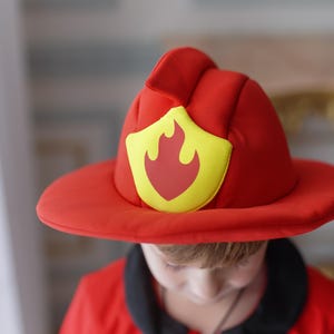May include: A child wearing a red firefighter costume with a yellow and red fire emblem on the hat. The costume has yellow stripes on the sleeves and a black collar.