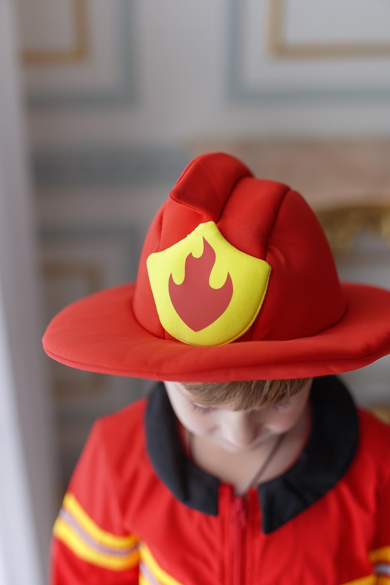 May include: A child wearing a red firefighter costume with a yellow and red fire emblem on the hat. The costume has yellow stripes on the sleeves and a black collar.