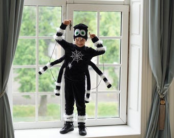 May include: A black spider costume featuring a long-sleeved top with a white spiderweb graphic, matching pants, and a beanie with large green and white eyes. The outfit includes four additional stuffed spider legs with white faux fur accents.