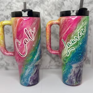 May include: Two personalized tumblers with handles, featuring a vibrant rainbow swirl design. One tumbler has the name "Cali" in white script, and the other has "Lauren" in green script. Both have black lids and straws.