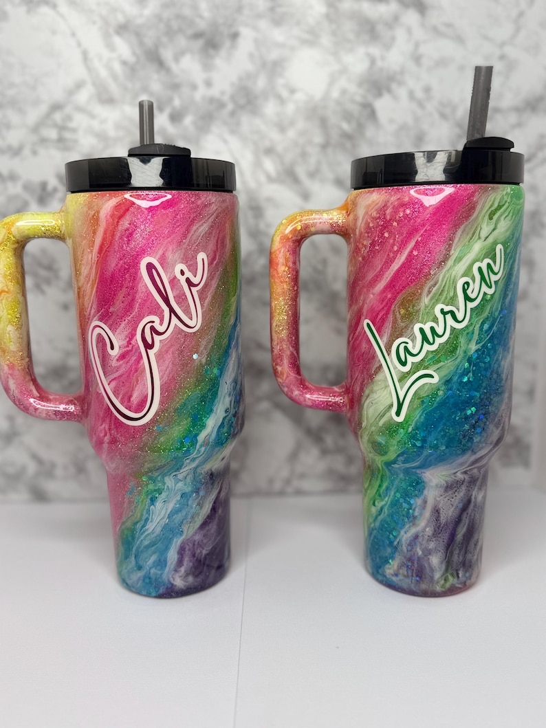 May include: Two personalized tumblers with handles, featuring a vibrant rainbow swirl design. One tumbler has the name "Cali" in white script, and the other has "Lauren" in green script. Both have black lids and straws.