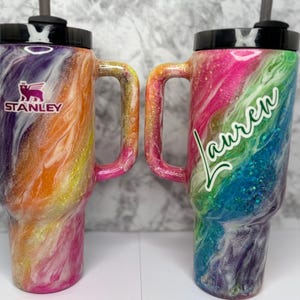 May include: Two colorful Stanley tumblers with handles and black lids. One features the Stanley logo, the other has the name "Lauren" in cursive. Both have a swirling design in shades of pink, orange, purple, and blue, with glitter accents.