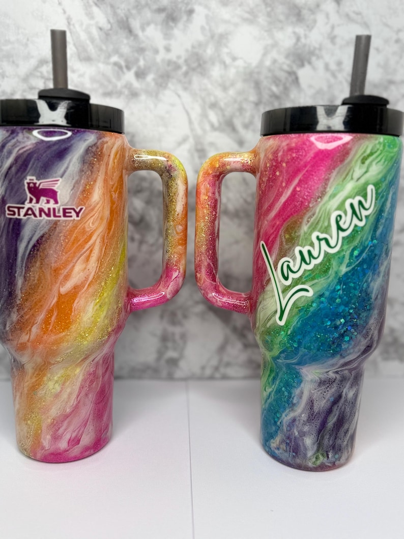 May include: Two colorful Stanley tumblers with handles and black lids. One features the Stanley logo, the other has the name "Lauren" in cursive. Both have a swirling design in shades of pink, orange, purple, and blue, with glitter accents.