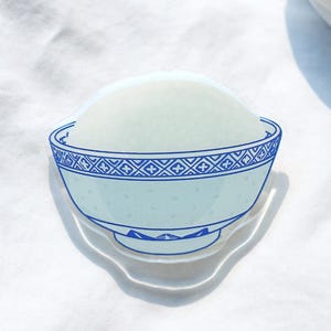 May include: A clear sticker featuring a bowl of rice with a blue and white decorative pattern. The sticker is on a white surface, with small, clear, ring-shaped embellishments scattered around it. A small potted succulent is in the background.
